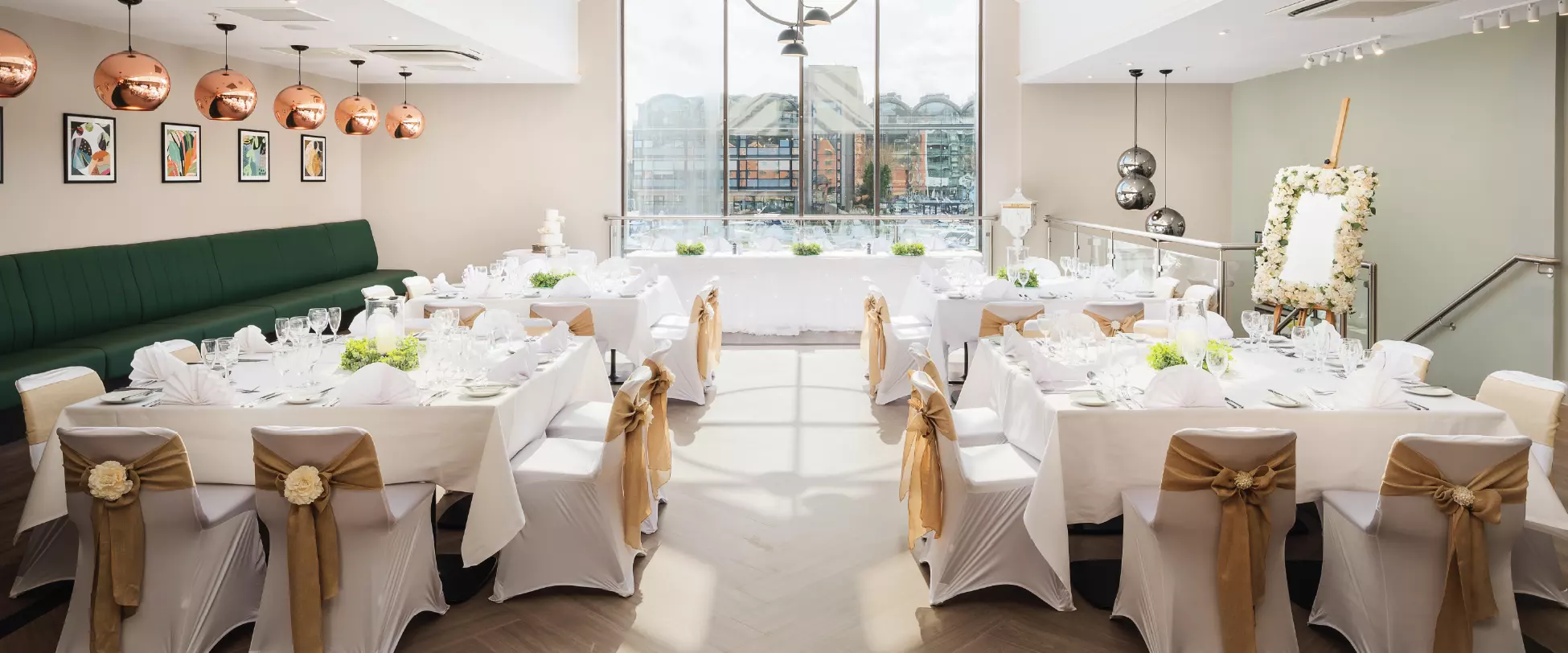 A large room set up for a wedding with tables and chairs covered in white, bronze ribbons and floral arrangements. There's a large window in the back overlooking a river..